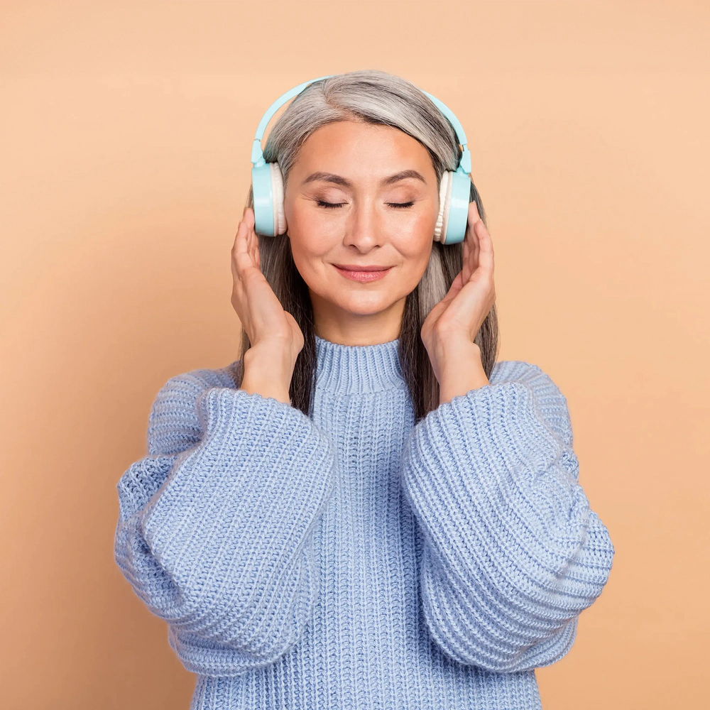 Kobieta w średnim wieku z siwymi włosami, zamniętymi oczami i nausznych słuchowakach na głowie, słuchająca muzyki trzymając na nich dłonie. Z lekkim uśmiechem na twarzy.