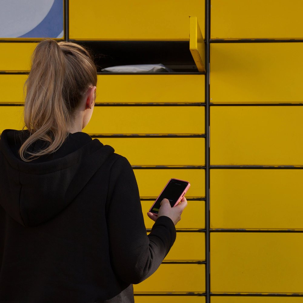 Młoda dziewczyna stojąca tyłem przed żółtym paczkomatem inpost, w czarnej bluzie z kitką, która trzyma w ręku telefon i otwiera jedną ze skrytek.
