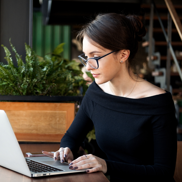 Młoda kobieta w ciemnych okularach i czarnej bluzce siedzi przy stoliku z otwartym laptopem.