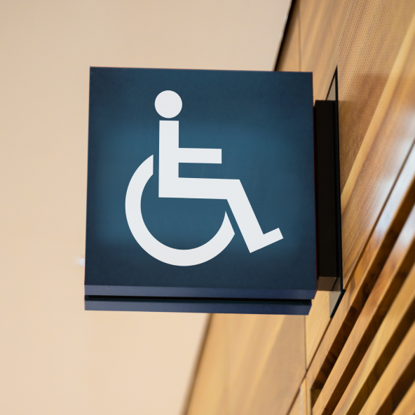 Czarna tabliczka z białą ikoną osoby niepełnosprawnej na wózku ortopedycznym, delikatnie podświetlana i zawieszona wysoko nad wejściem do toalet.