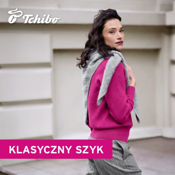 Kobieta w różowym swetrze, szarych spodniach w prążki i z szarą dzianiną przewieszoną przez ramiona. W tle jasna fasada budynku. Na grafice logo Tchibo i napis „Klasyczny szyk”.